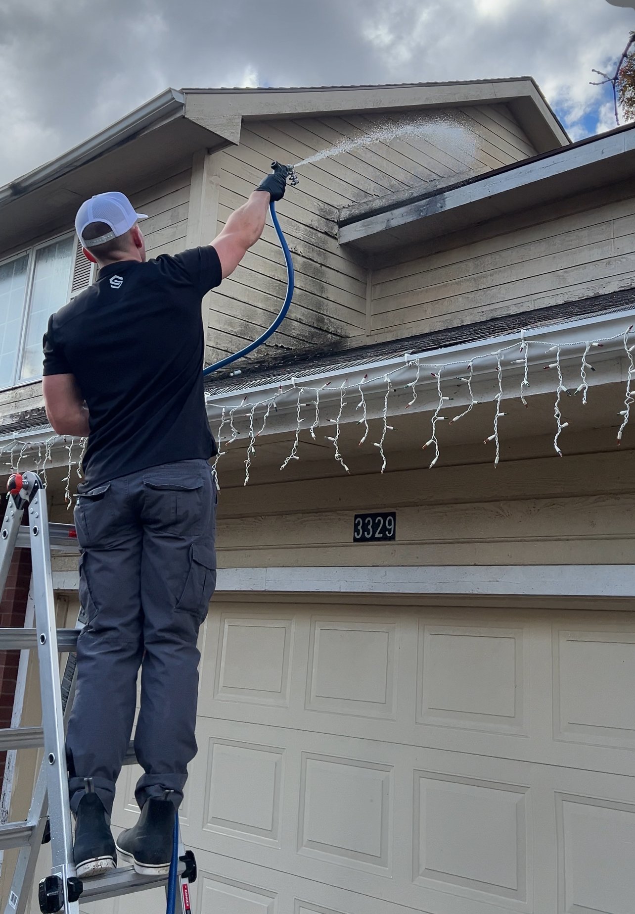 A man standing on a ladder power washing the exterior of a two-story house, located above a garage, with Christmas lights hanging from the eaves.