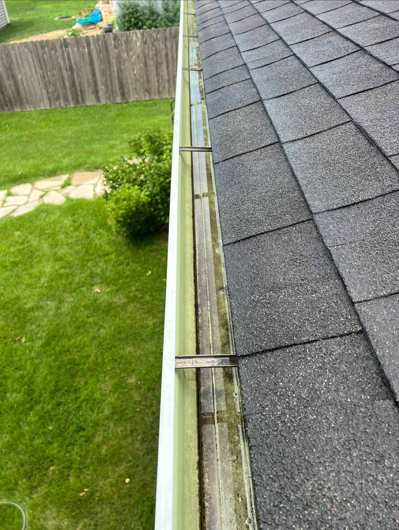 Close-up of roof gutter with some debris, alongside asphalt shingle roof, with green lawn and garden visible below.