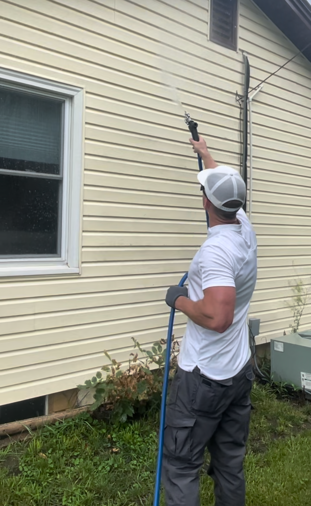 A man wearing a white cap, gray gloves, and a white t-shirt is pressure washing the exterior wall of a house with beige vinyl siding. He is holding a pressure washer nozzle and spraying water upward. There is a window with a white frame on his left, 