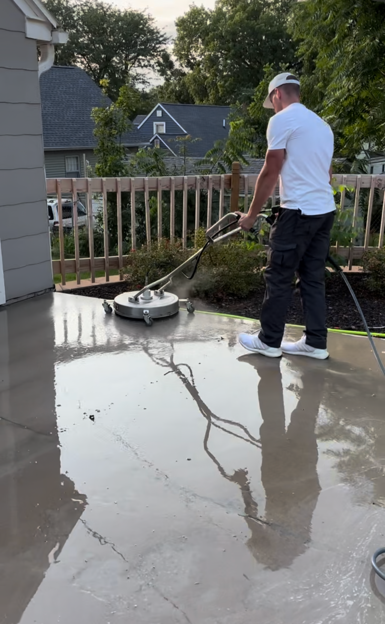 A man is pressure washing a concrete patio outside a house, with water spraying on the surface and wet reflections visible.