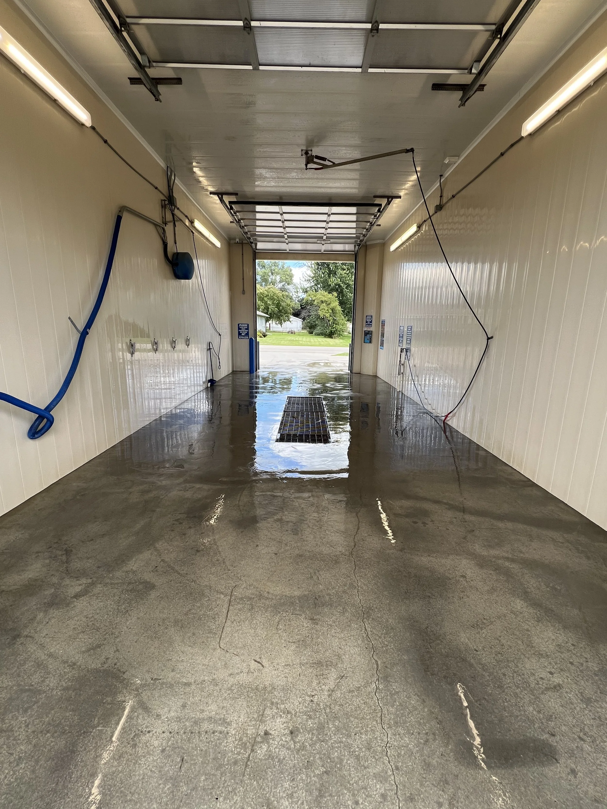 Empty car wash bay with wet concrete floor, hoses and equipment on the walls, open garage door showing trees outside, and a drainage grate in the middle of the floor.