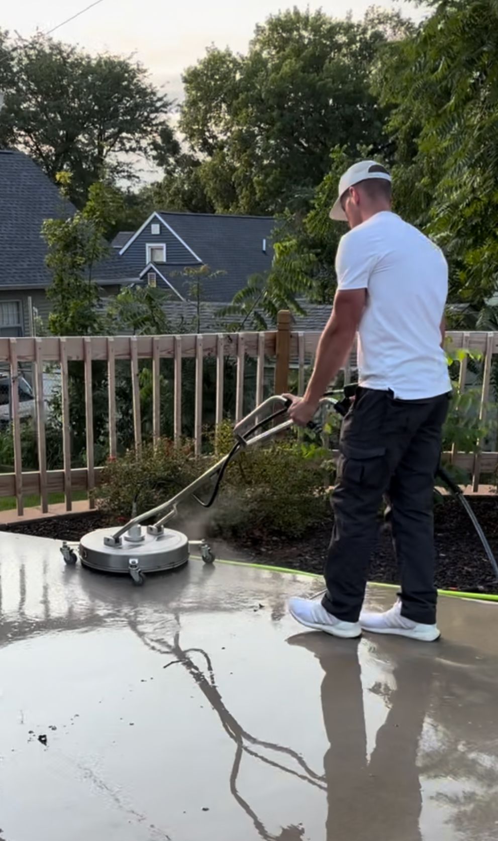 Man wearing white shirt, black pants, and a cap using a power scrubber on a wet concrete patio outdoors during daytime, with trees and houses in the background.