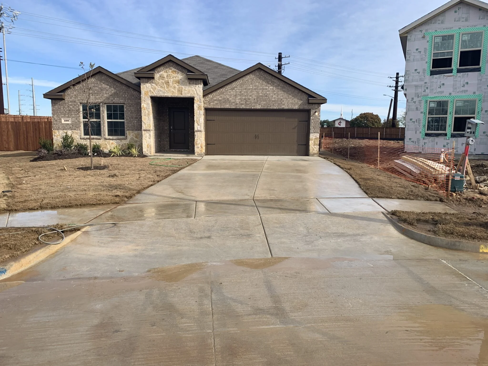 Newly constructed house with a two-car garage and stone accents, front yard with sparse grass and a young tree, concrete driveway, and sidewalk in progress, construction materials on the right side, and a neighboring house under construction.