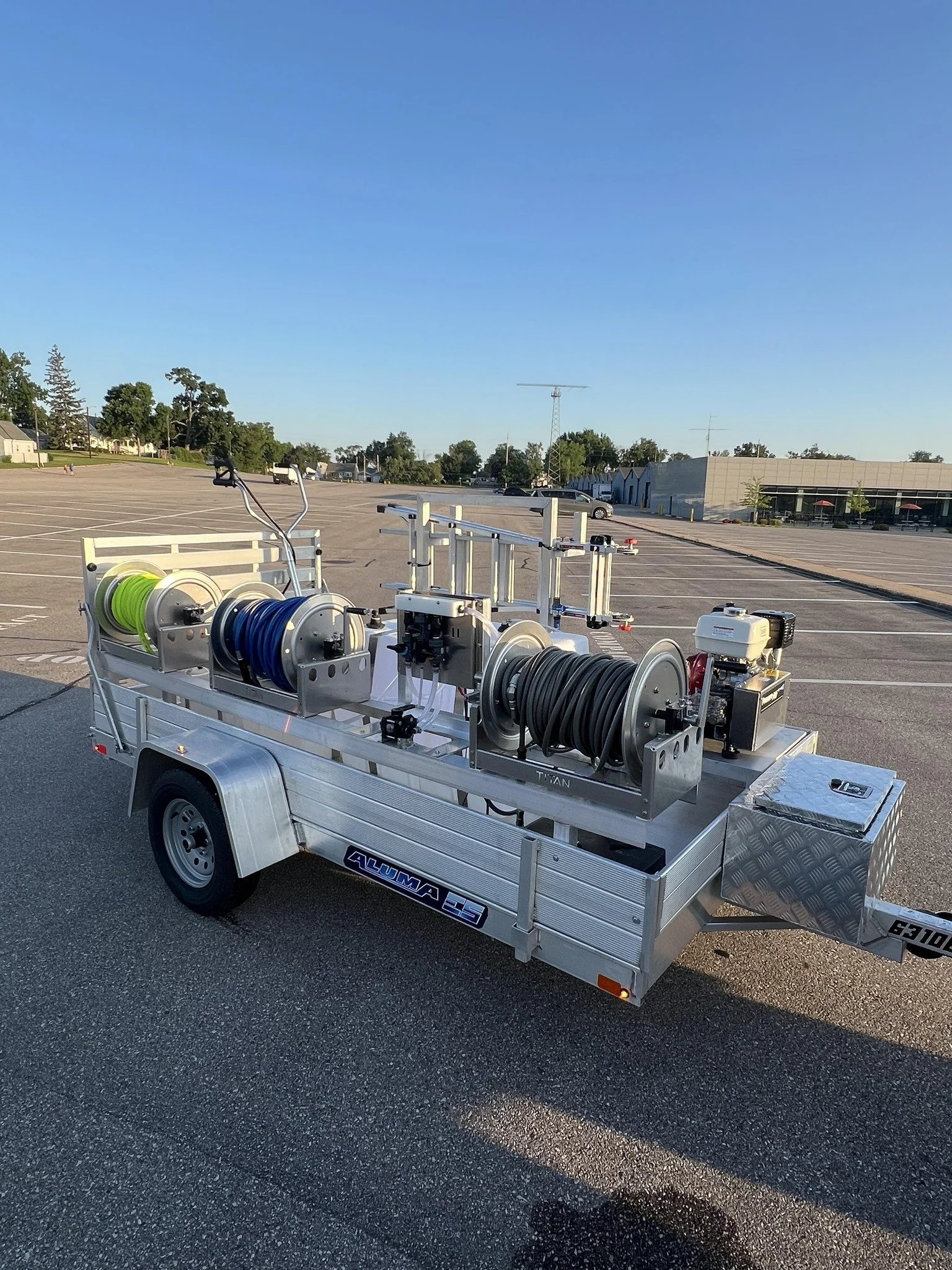 A utility trailer with hose reels and various equipment, parked in an empty parking lot under a clear blue sky.
