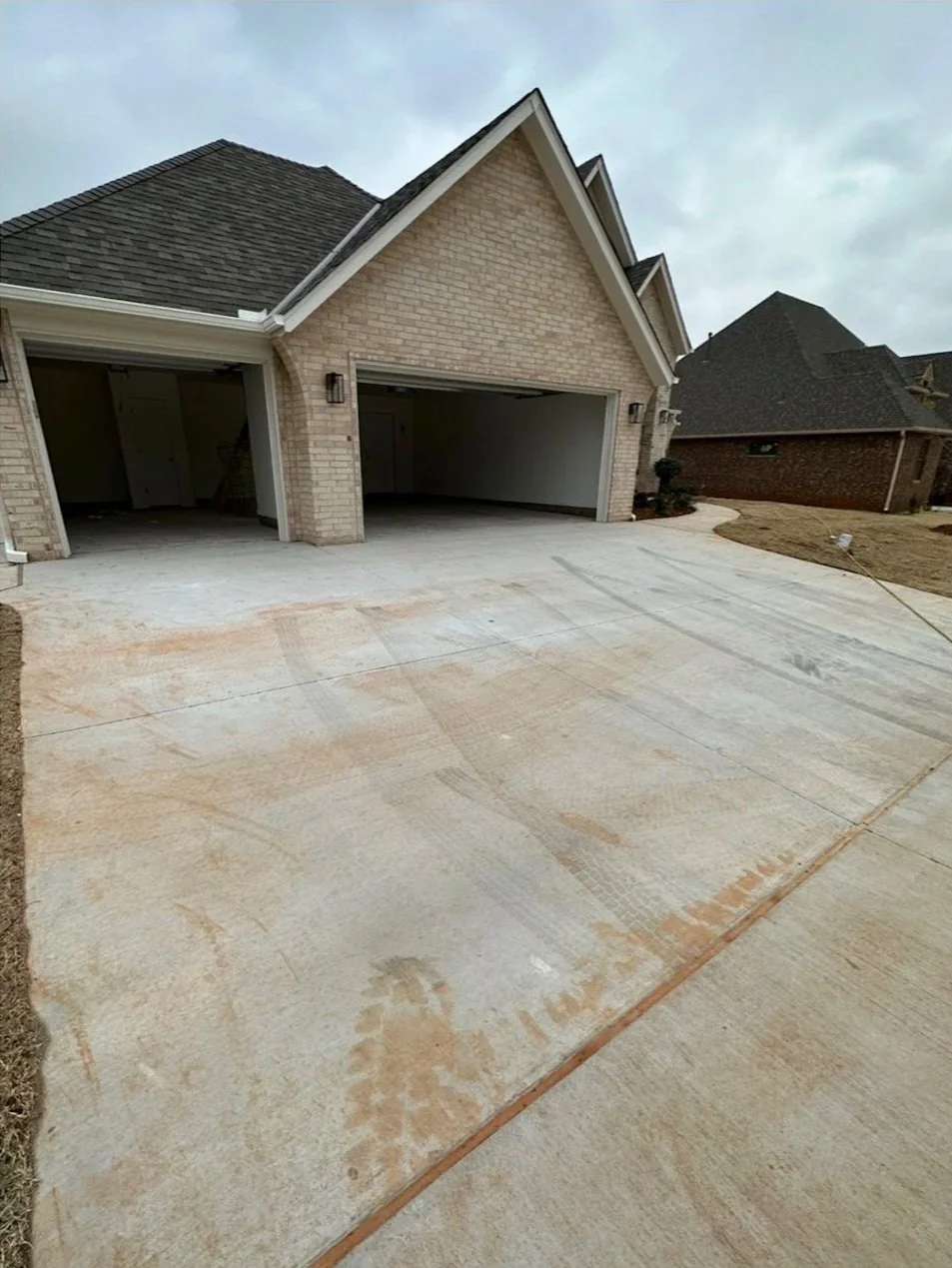 Newly constructed house with a unfinished concrete driveway and brick exterior, two open garage doors, and neighboring houses visible under an overcast sky.