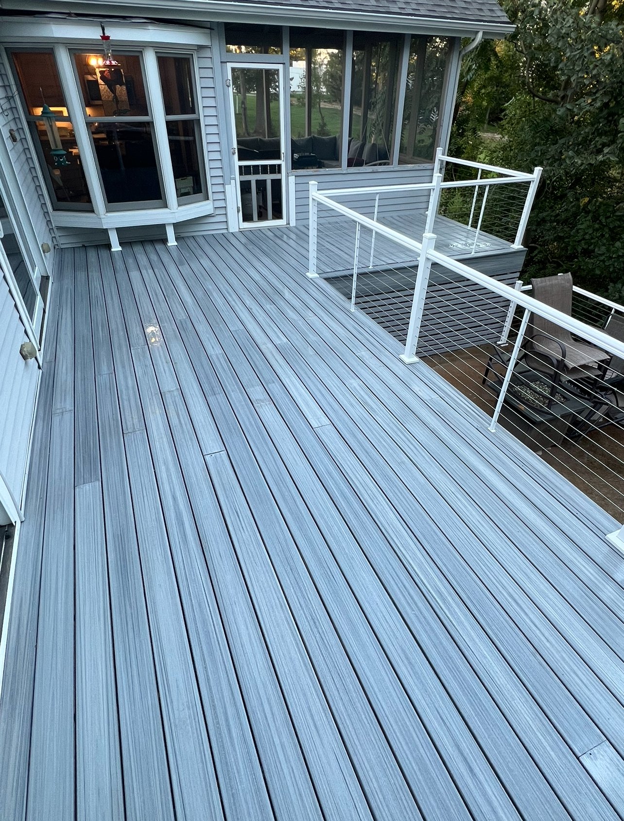 Empty outdoor deck with light gray wooden planks, white railing, attached to a house with glass sliding doors and windows, and a screened-in porch area. Trees and green grass are visible in the background.