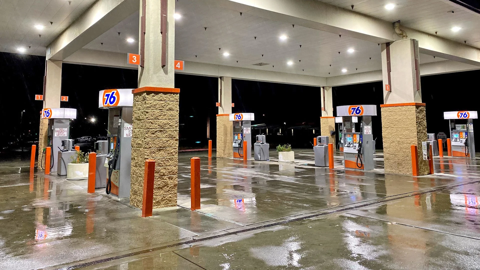 Empty gas station with wet pavement and no cars at the pumps, illuminated by overhead lights at night.