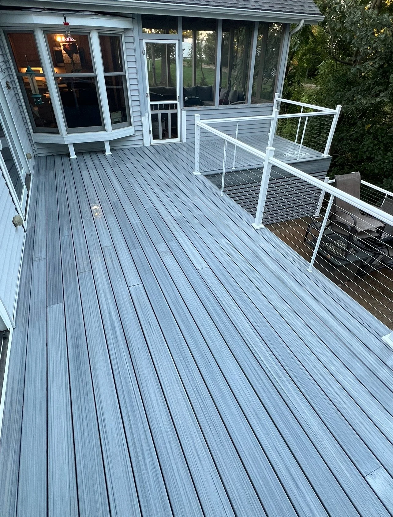 A spacious outdoor deck with grey composite flooring, surrounded by white metal railing, attached to a house with large windows and a screened porch, with outdoor furniture visible under the trees.