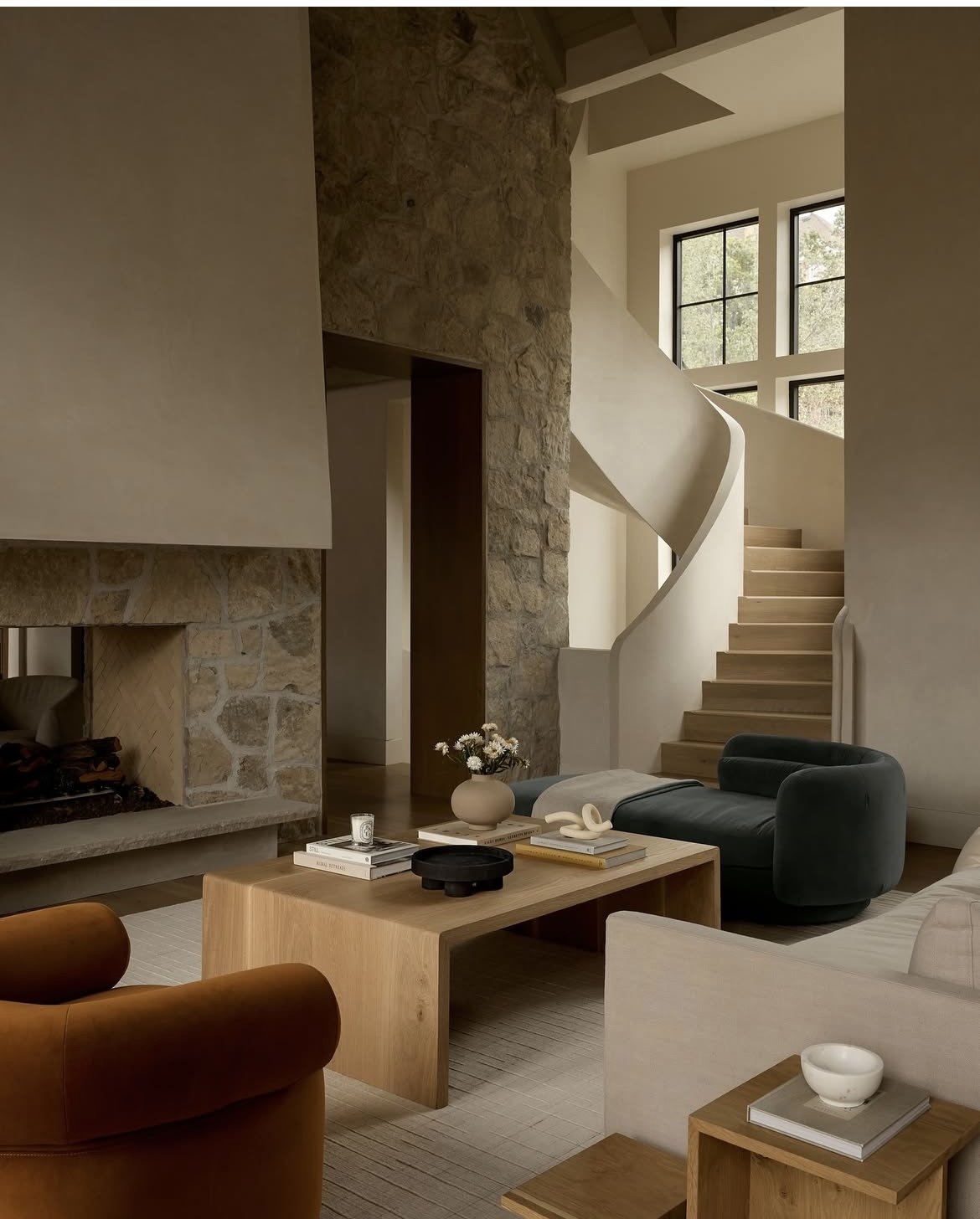 Modern living room with stone fireplace, wooden furniture, spiral staircase, and large windows. Chic decor includes vases, books, and a cozy sofa. Custom furniture design High-end furniture manufacturing Bespoke furniture American-made furniture.