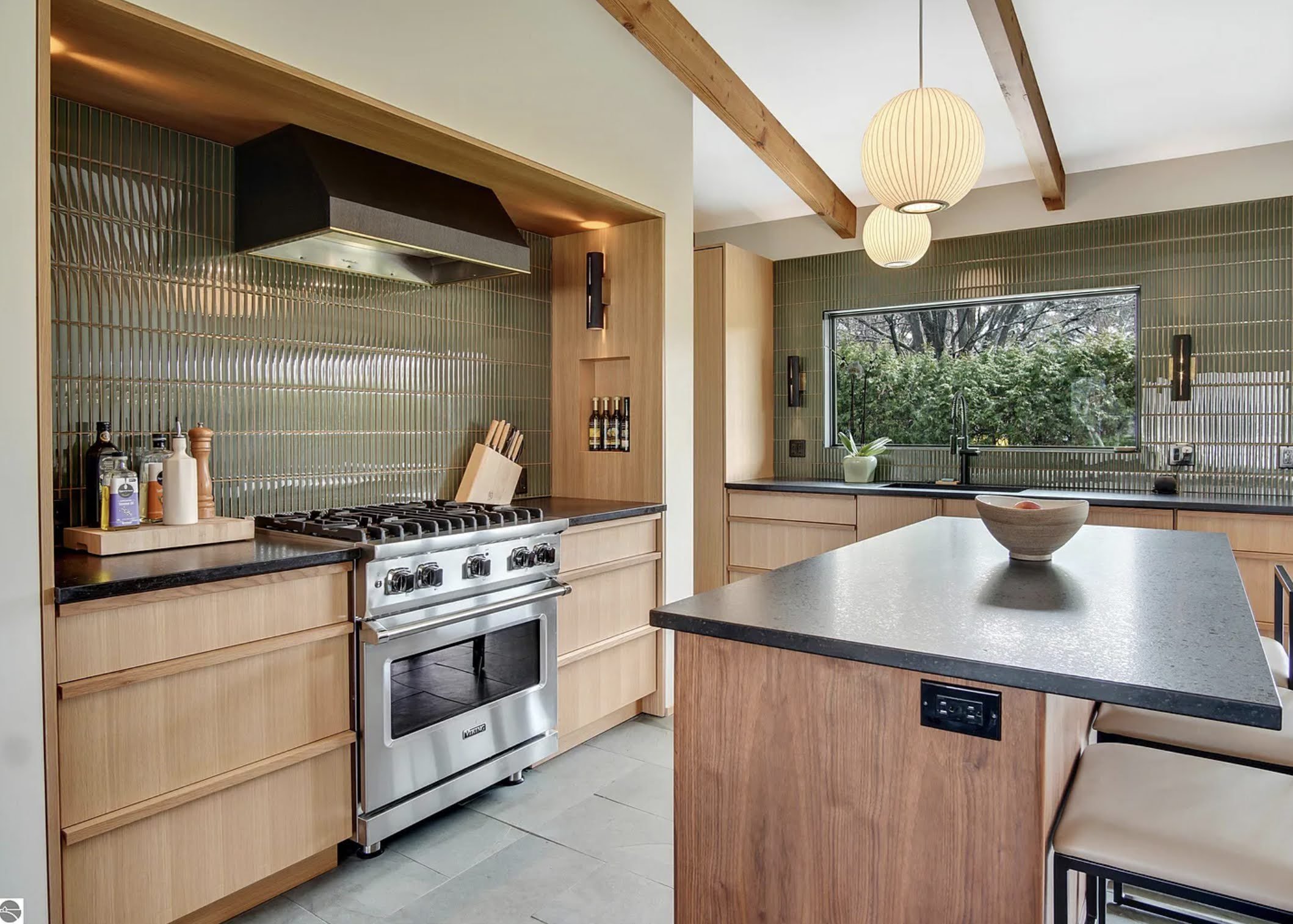 Modern kitchen with stainless steel stove, wood cabinetry, green tile backsplash, and pendant lighting. Large window above sink, featuring outdoor view.