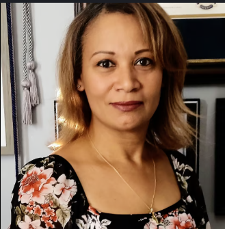 A woman with shoulder-length, light brown hair taking a selfie indoors, wearing a floral top and a gold necklace.