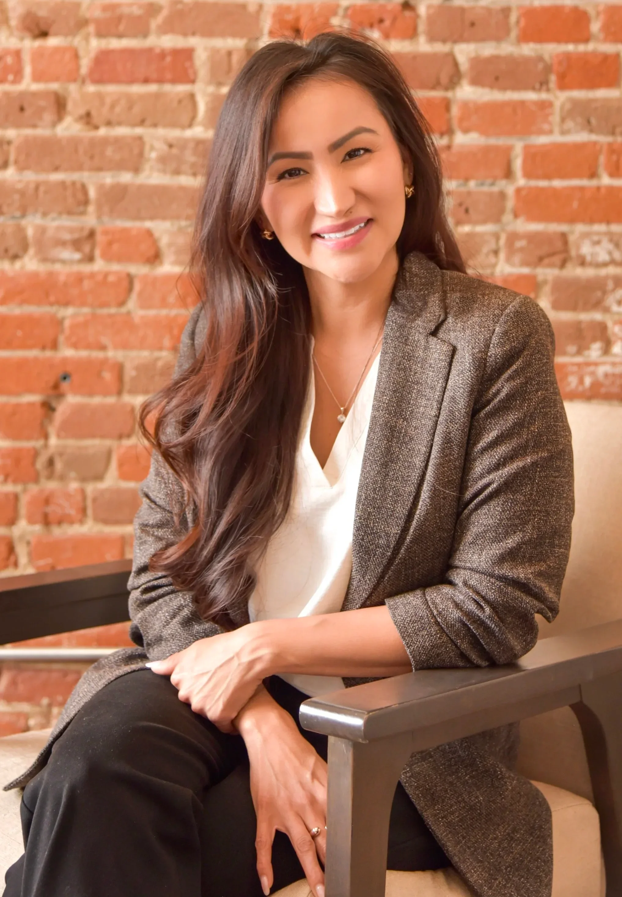 A woman with long, wavy brown hair sitting on a beige chair in front of a red brick wall, smiling at the camera, wearing a brown blazer over a white top, and black pants.