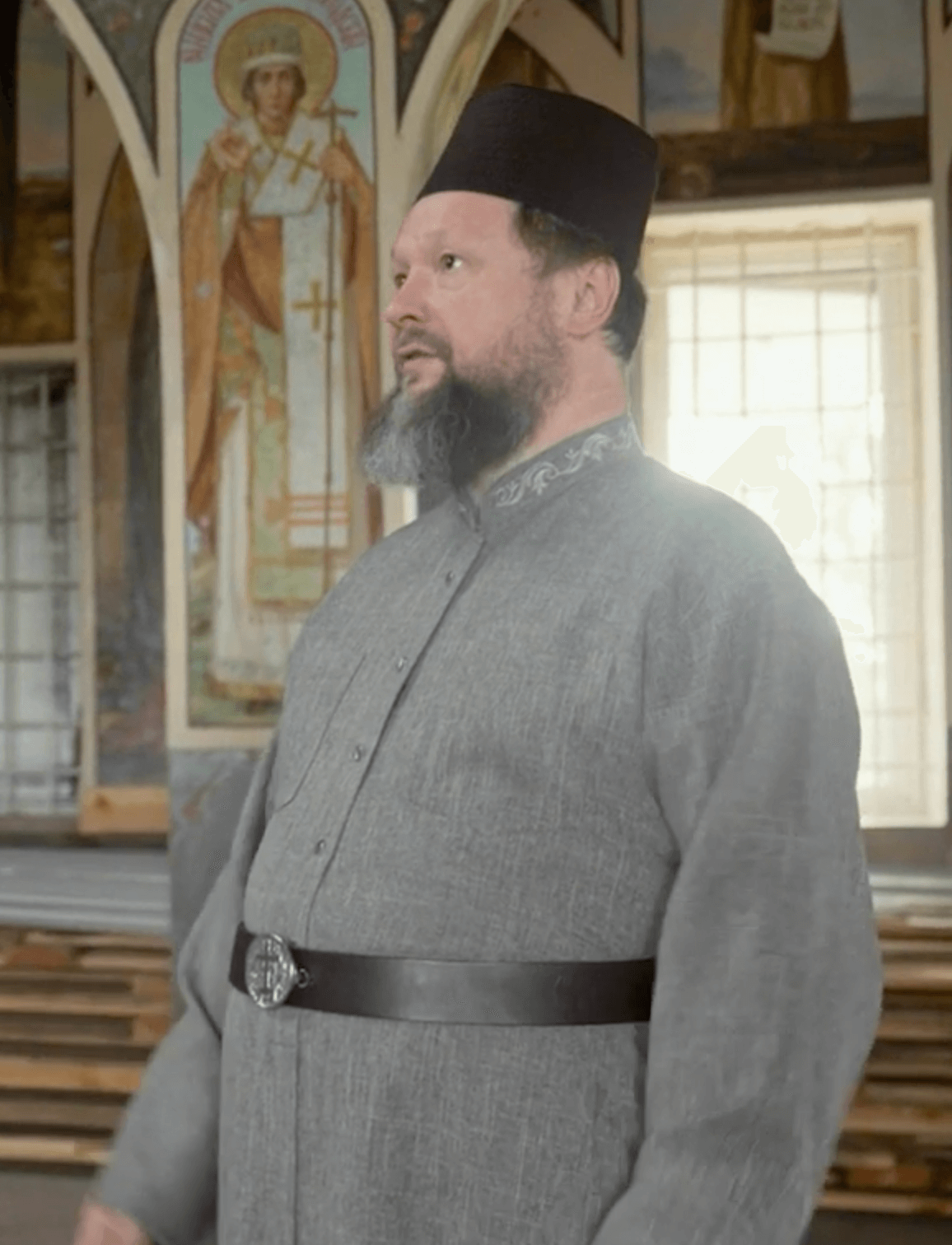 A man dressed in traditional Eastern Orthodox clerical attire, wearing a black kalimavkion and gray cassock, standing inside a church with religious icons and icons in the background.