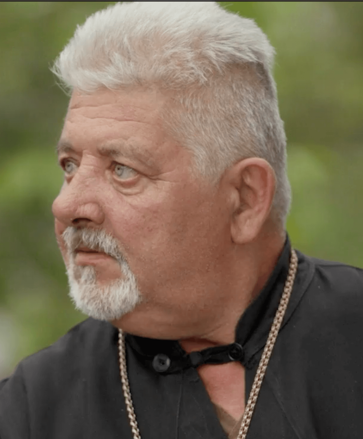 Close-up of an older man with a white goatee and mustache, gray hair, wearing a black shirt with a collar and a gold chain, outdoors with a blurred green background.