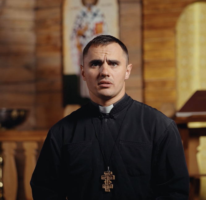 A man dressed as a priest with a surprised or confused expression, standing inside a church or chapel with wooden walls and religious decor in the background.