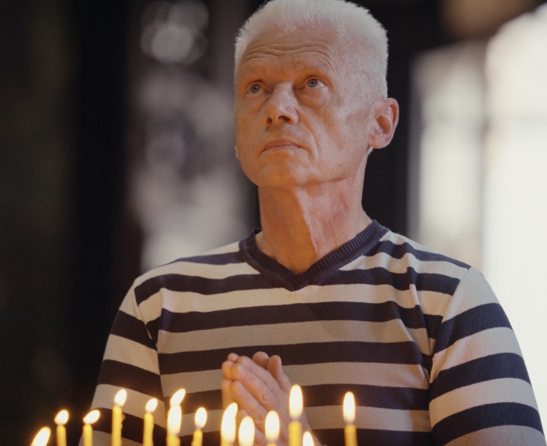 Elderly man with white hair and striped shirt looking upward with candles in front.