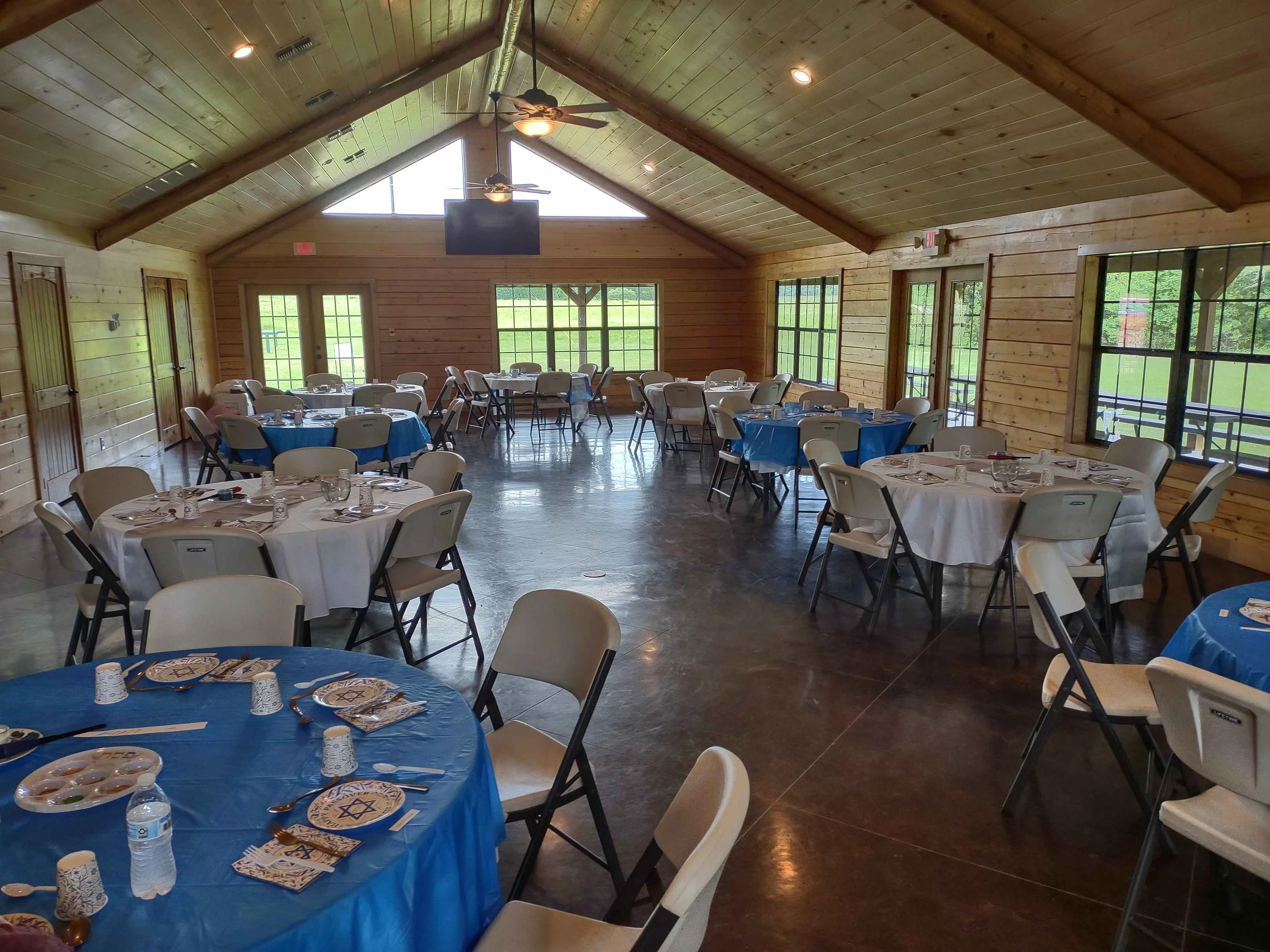 Indoor event space with wooden walls and ceiling, decorated for a celebration. Round tables with blue tablecloths, paper plates, napkins, plastic utensils, and small cups are set up.