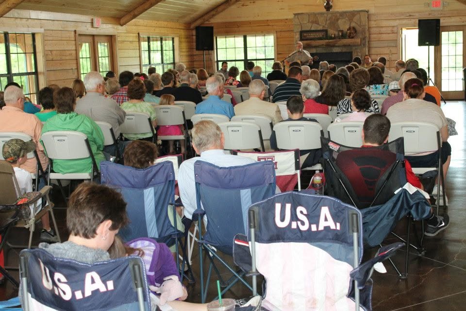People attending a gathering in a hall, sitting on chairs and blankets, listening to a speaker near a fireplace.