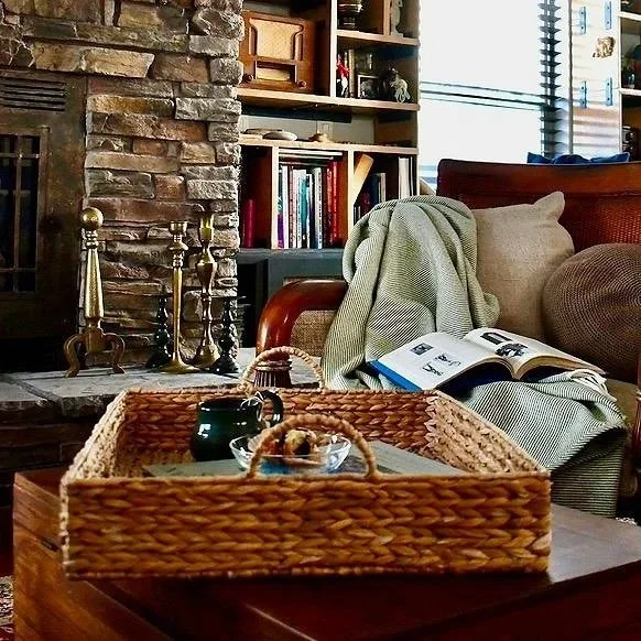 Cozy living room with a stone fireplace, a shelf filled with books, a cushioned armchair with a folded blanket, a wicker tray on a wooden table holding a black mug, a glass bowl with snacks, and an open magazine.