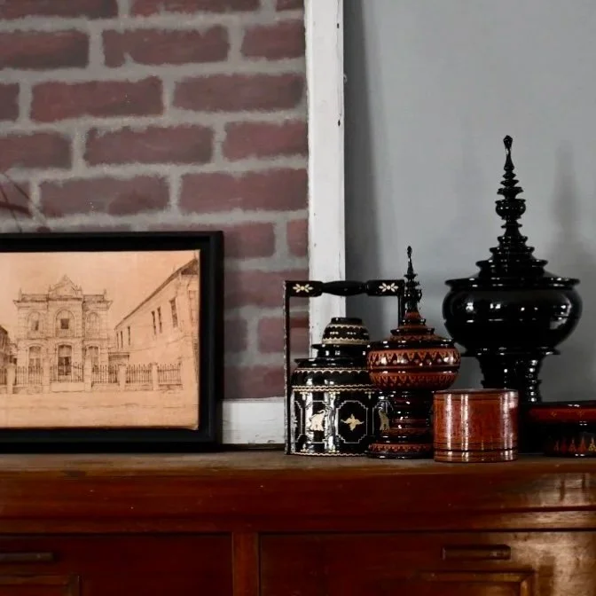 Decorative Thai-style containers and a framed sketch of a building on a wooden surface against a brick wall and a white wall.