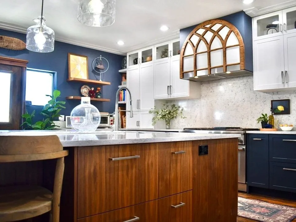 Modern kitchen with blue walls, white cabinets, and a wooden kitchen island. Decor includes glass pendant lights, a vase, shelves with decorative items, and a small plant. There is a window with a plant nearby and various kitchen appliances.