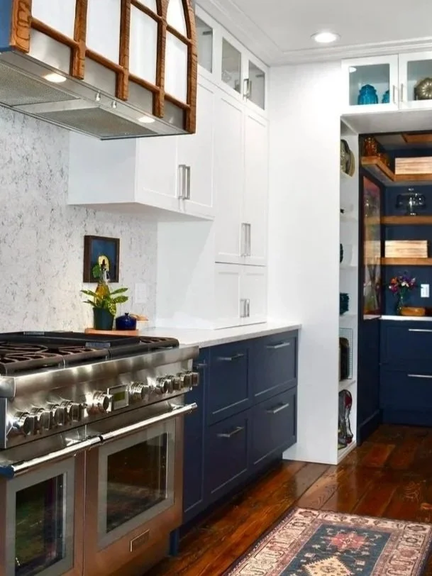 A modern kitchen with white upper cabinets and blue lower cabinets, a stainless steel oven and stove, a marble backsplash, a small potted plant on the counter, open shelving above with wooden racks, and a wooden floor with a decorative rug.