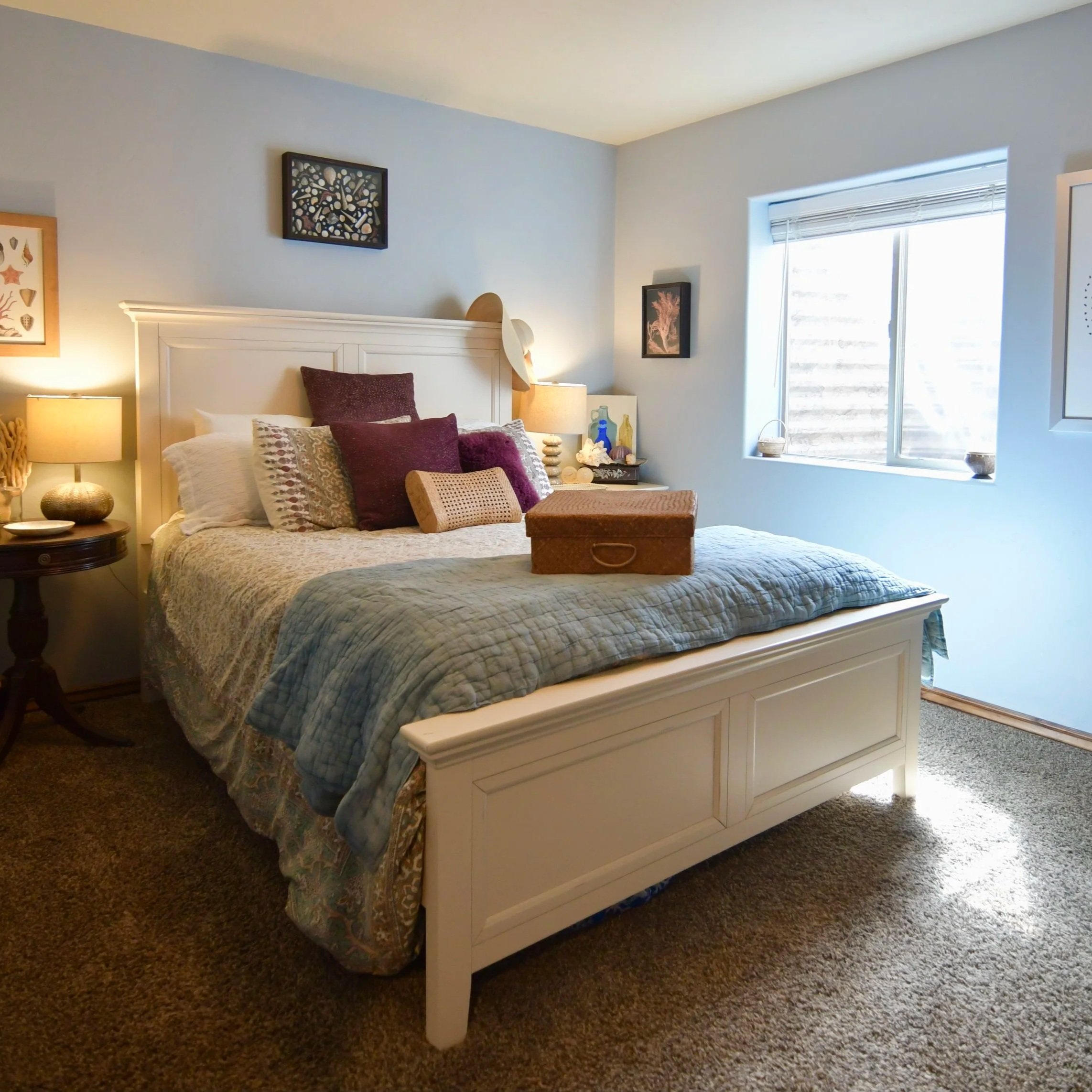 A neatly made bedroom with a white wooden bed frame, various purple and patterned pillows, a brown quilt, and a wicker suitcase on top. There are bedside lamps, framed art pieces on the walls, a window with blinds, and a small circular table with decor items.