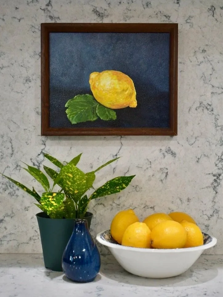 A framed painting of a lemon with green leaves hangs on a marble wall, a potted plant with green and yellow variegated leaves, a small blue vase, and a white bowl filled with lemons are placed on a marble surface.
