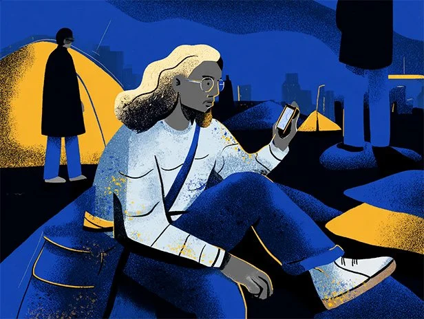 A person sitting on the ground in a tent encampment looks at their phone, trying to access services. They have long hair, glasses, and an ambiguous gender presentation.