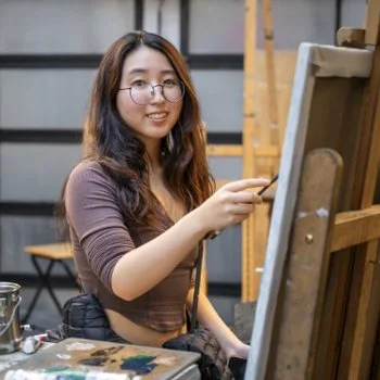 A young woman smiles with a hand raised—paintbrush in hand—painting an easel-mounted canvas in studio.