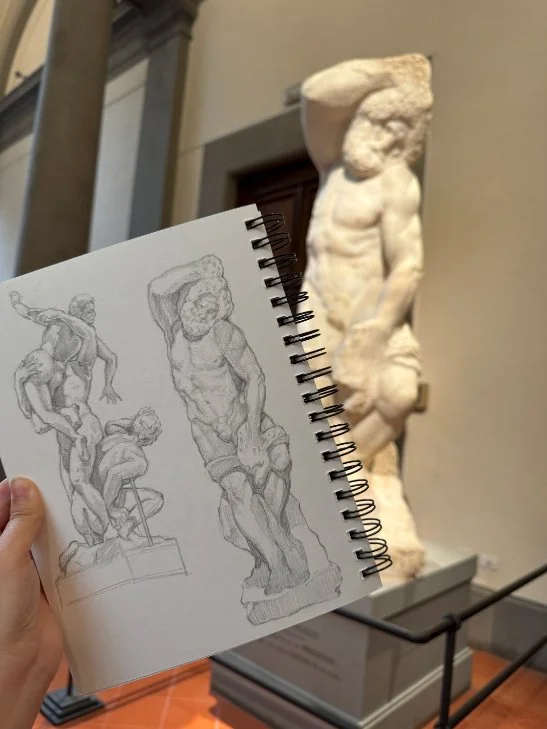 A hand holds up a couple of sketches of sculptures, behind the pencil sketches one of the featured sculptures is lit in museum lights.