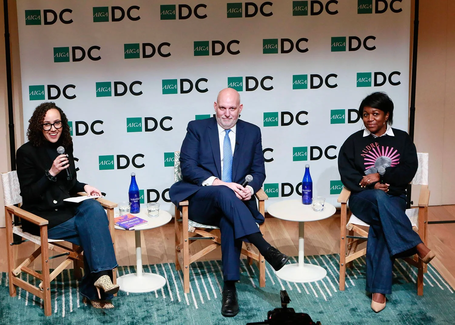 Ashleigh Axios moderates a panel discussion with two speakers at an AIGA DC event, seated on stage in conversation with microphones and small tables during a public dialogue on design and leadership.