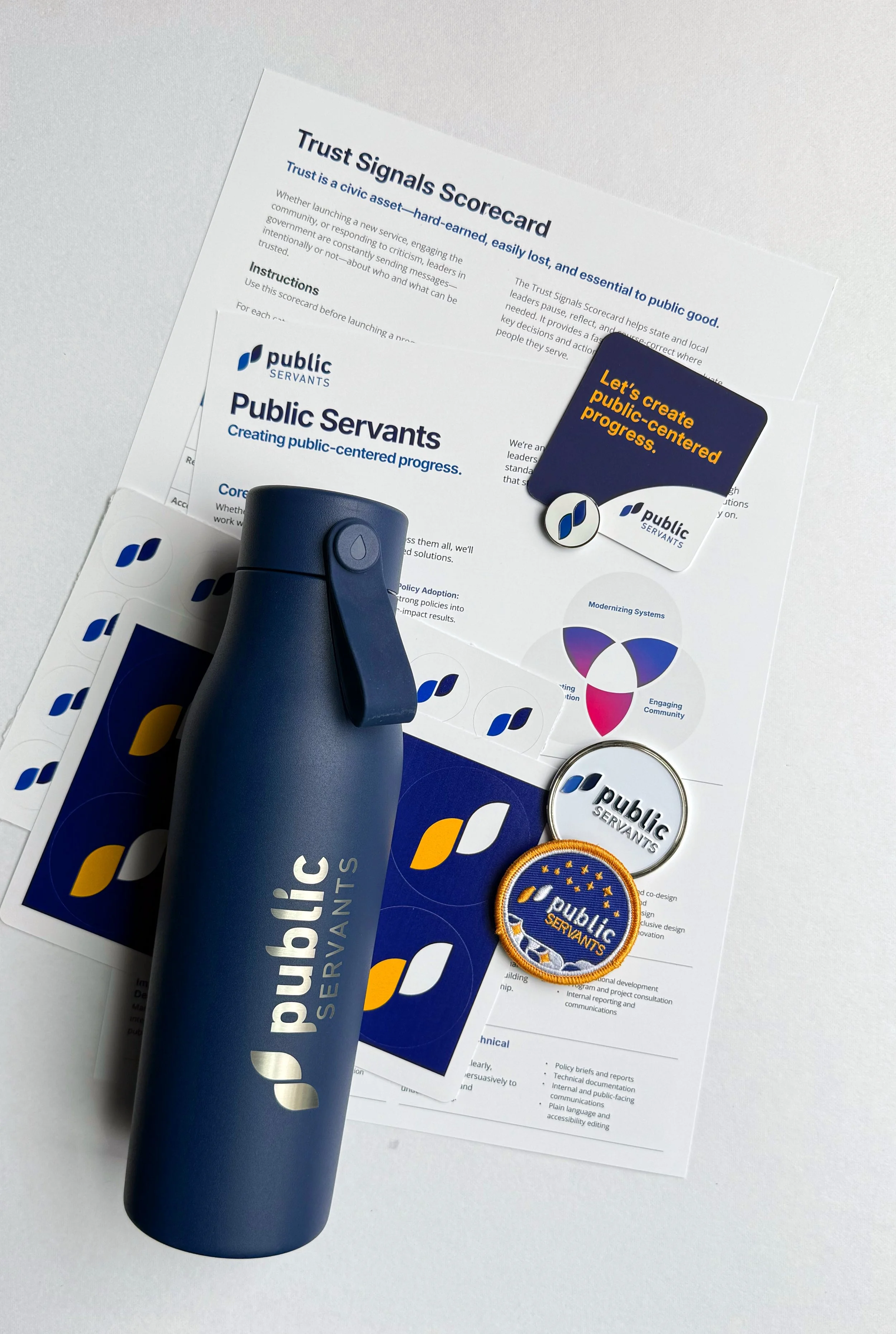 Flat lay of Public Servants materials including the Trust Signals Scorecard, framework cards, stickers, and a branded water bottle, representing tools used in public-centered design work.