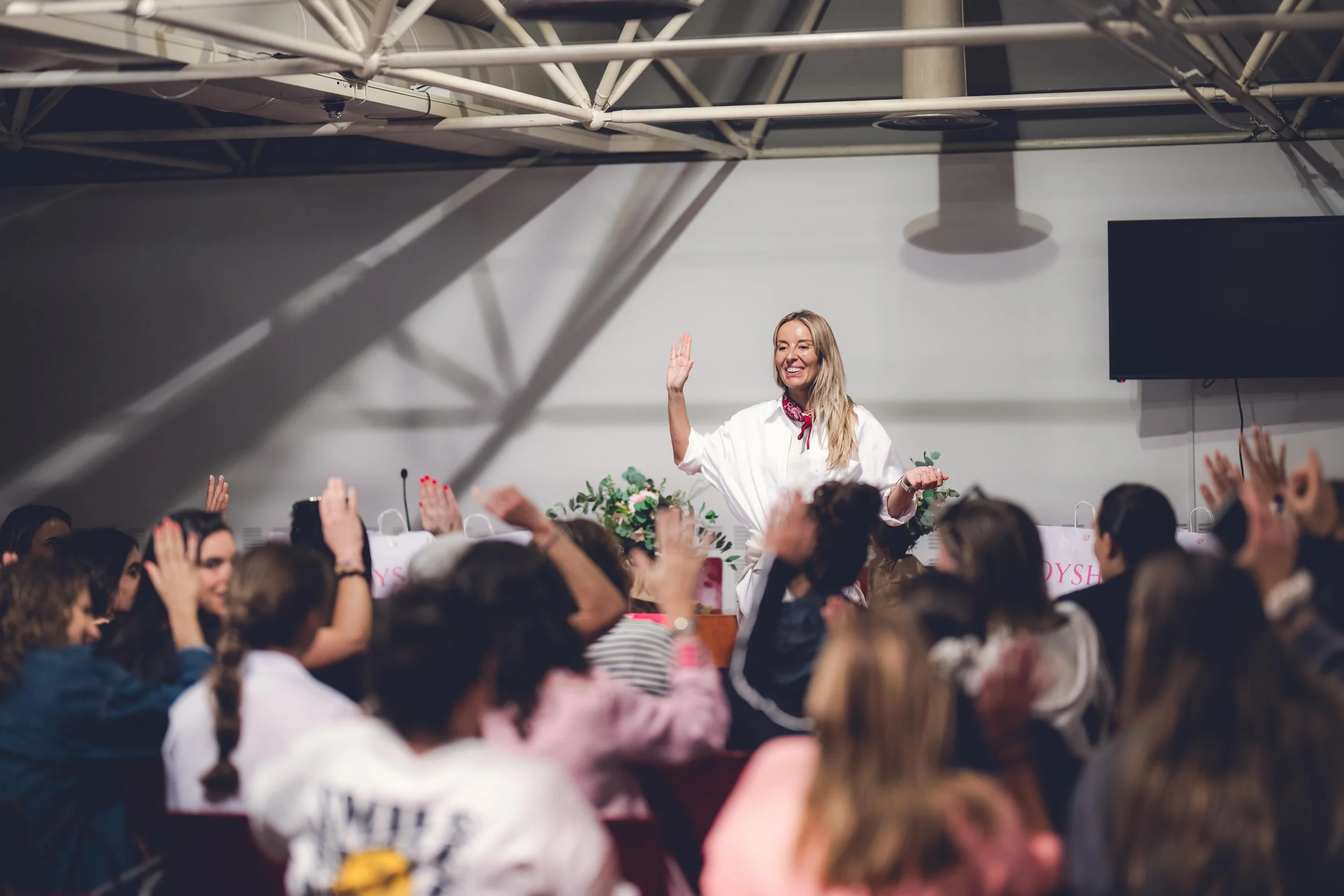 Mujer en el escenario saludando a la audiencia en un evento, rodeada de muchas personas que levantan sus manos en señal de participación o saludo.