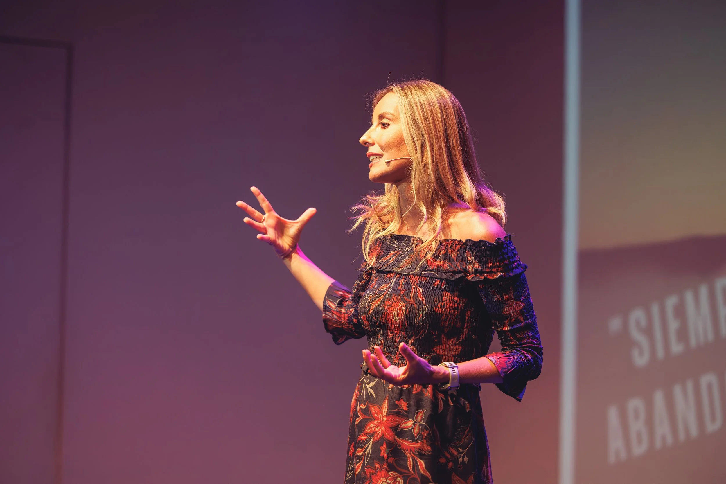 Una mujer sonriente con vestido largo y abierto en la parte frontal, levantando los brazos en señal de celebración o victoria, en un escenario con mesa y sillas.