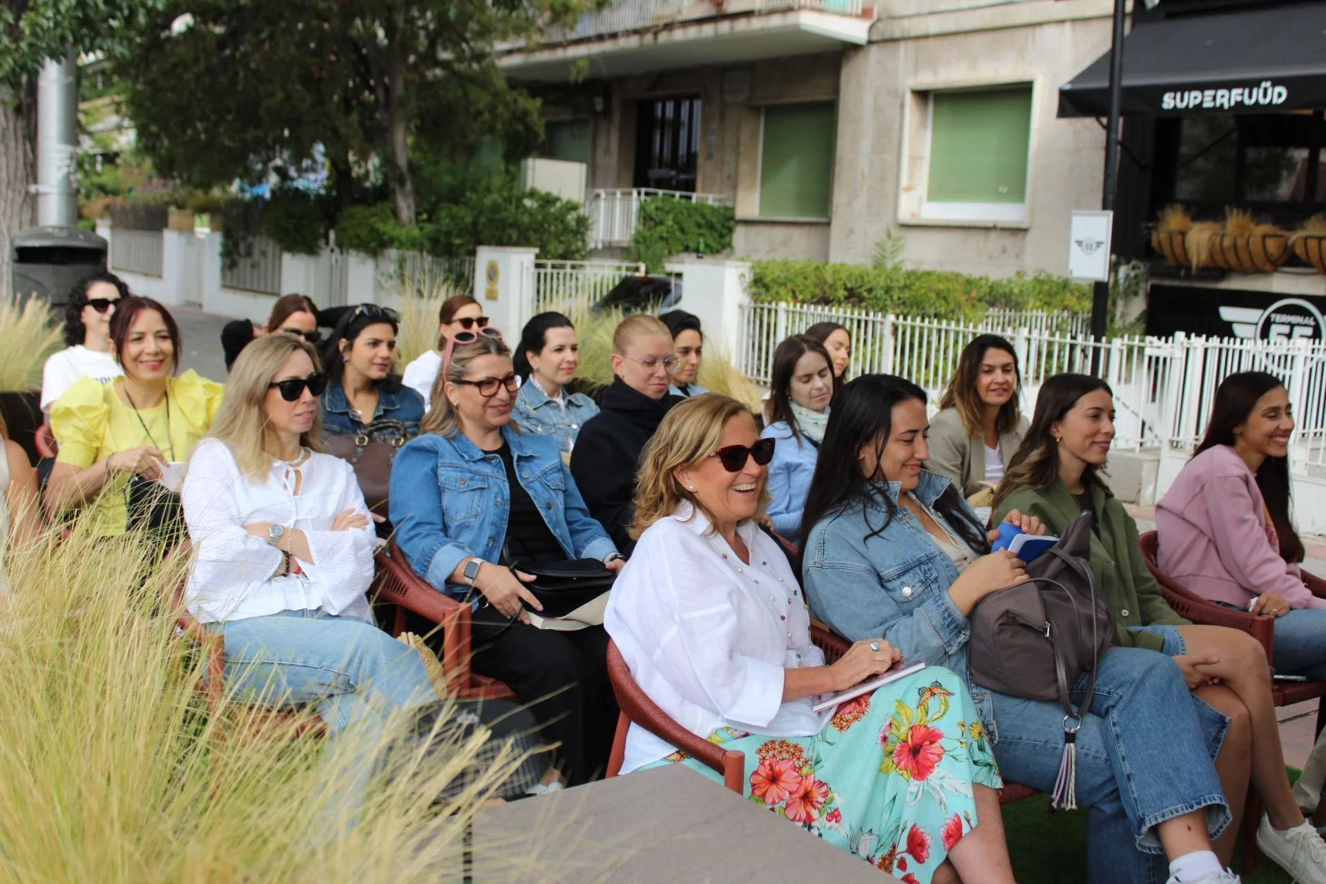Grupo de mujeres sentadas en una reunión al aire libre, algunos tomando notas y otros sonriendo, con fondo de edificio y vegetación.
