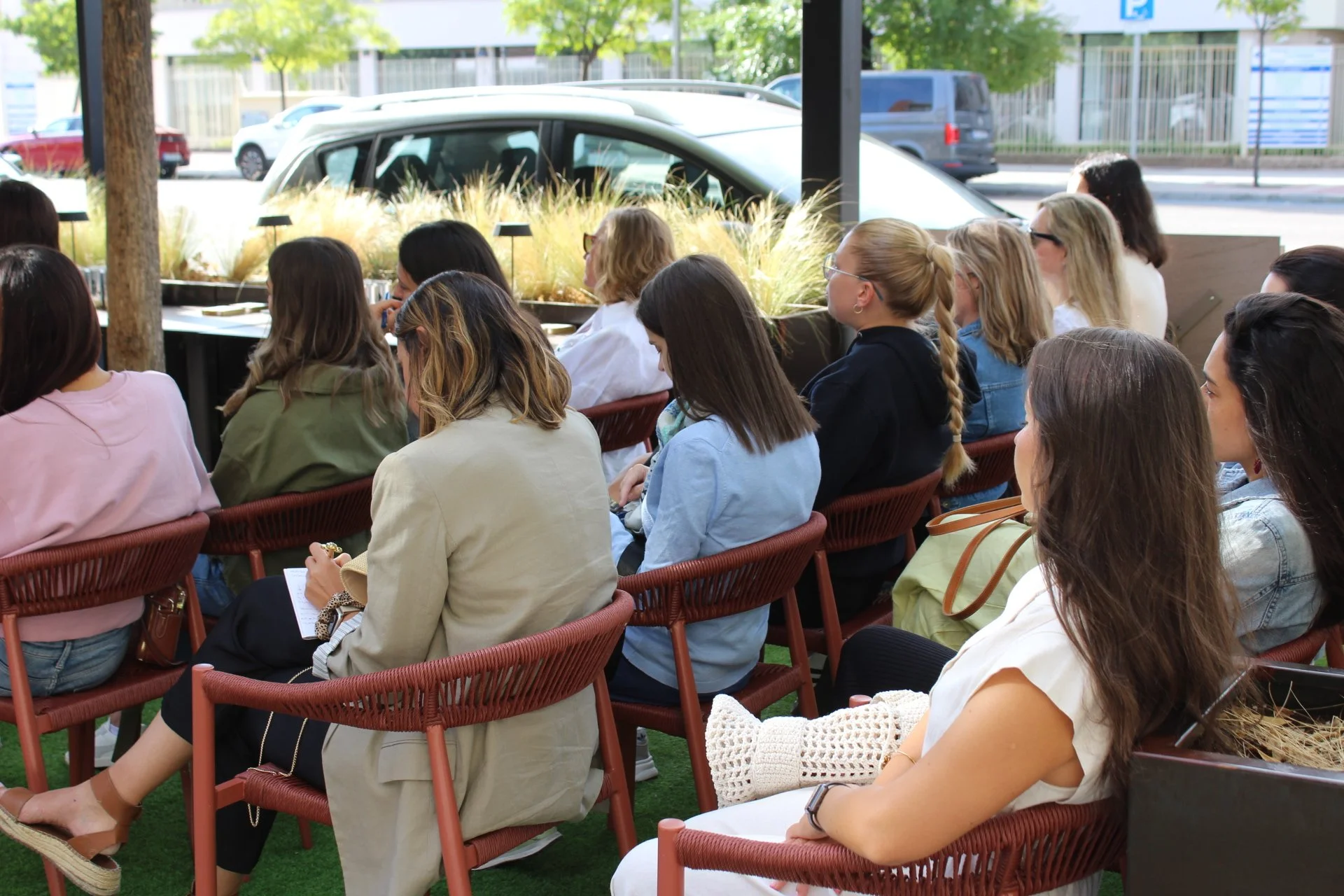 Grupo de mujeres sentadas en sillas en un evento, en un espacio interior con vista al exterior y coches estacionados.