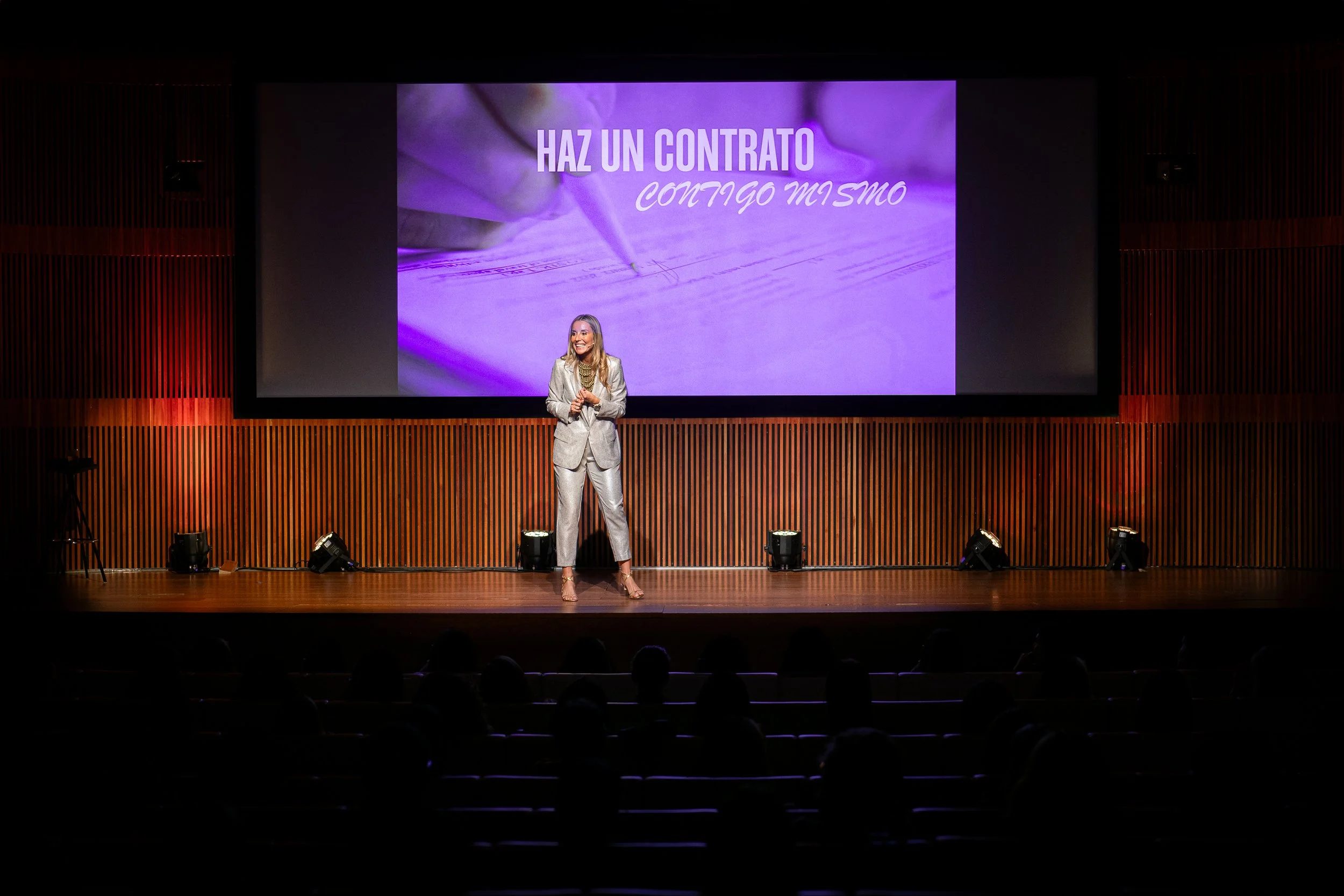 Una mujer vestida con traje plateado presenta en un escenario con una pantalla que dice 'Haz un contrato contigo mismo', en un auditorio con público sentado en la oscuridad.