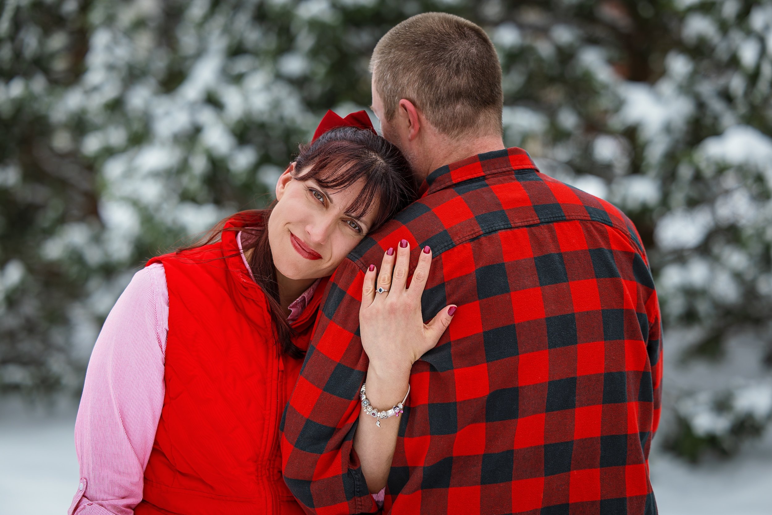 Woman facing forward showing her ring hand with engagement ring, leaning on her fiancé facing away from the camera