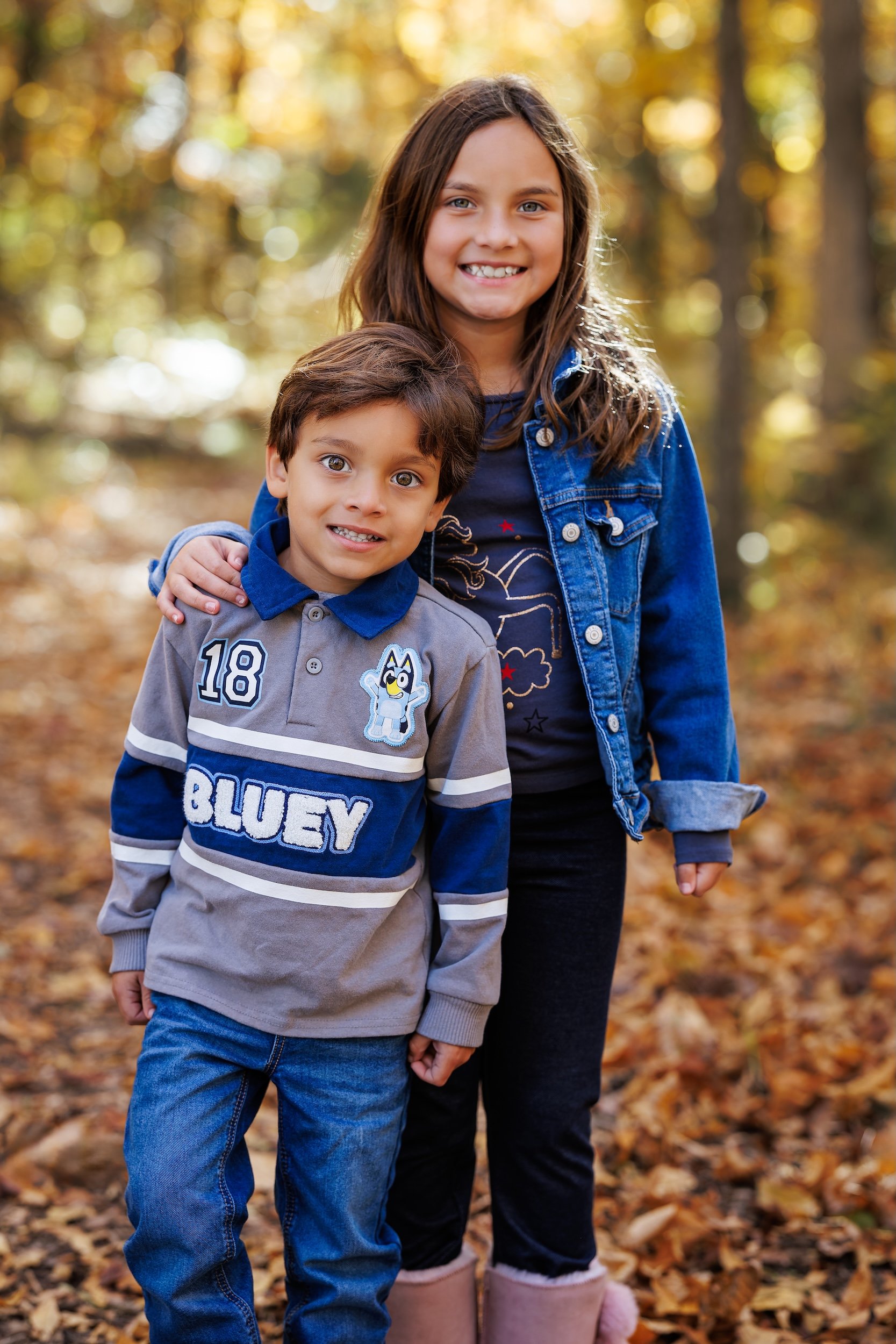 Older sister posing with her younger brother, both smiling with fall foliage in the background