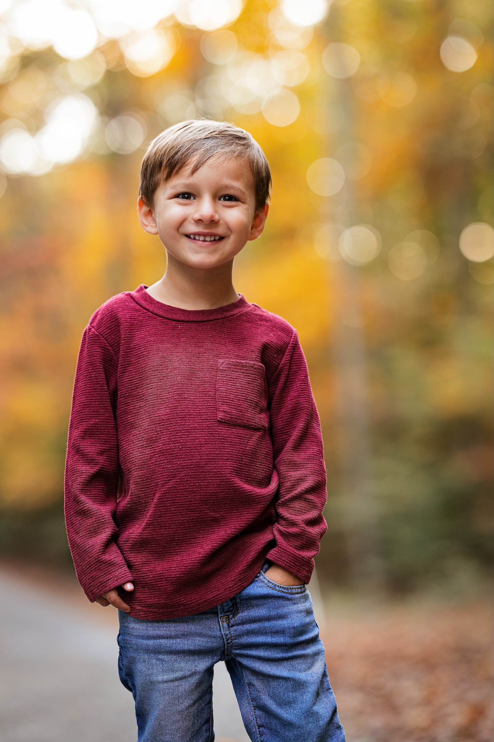 Young boy smiling with a red shirt and jeans with fall foliage in the background
