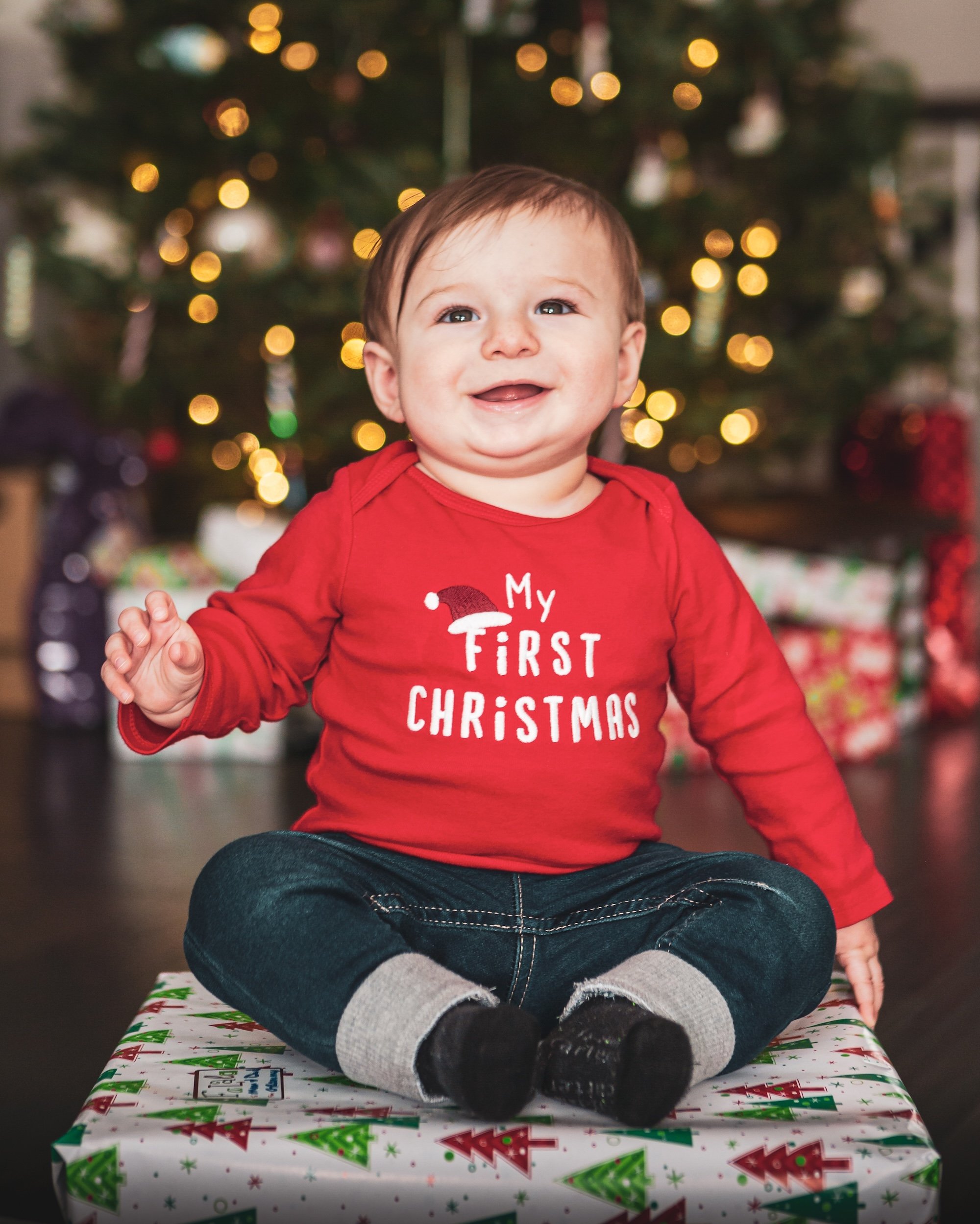 Baby smiling at the camera wearing festive outfit with Christmas tree lit up in the background