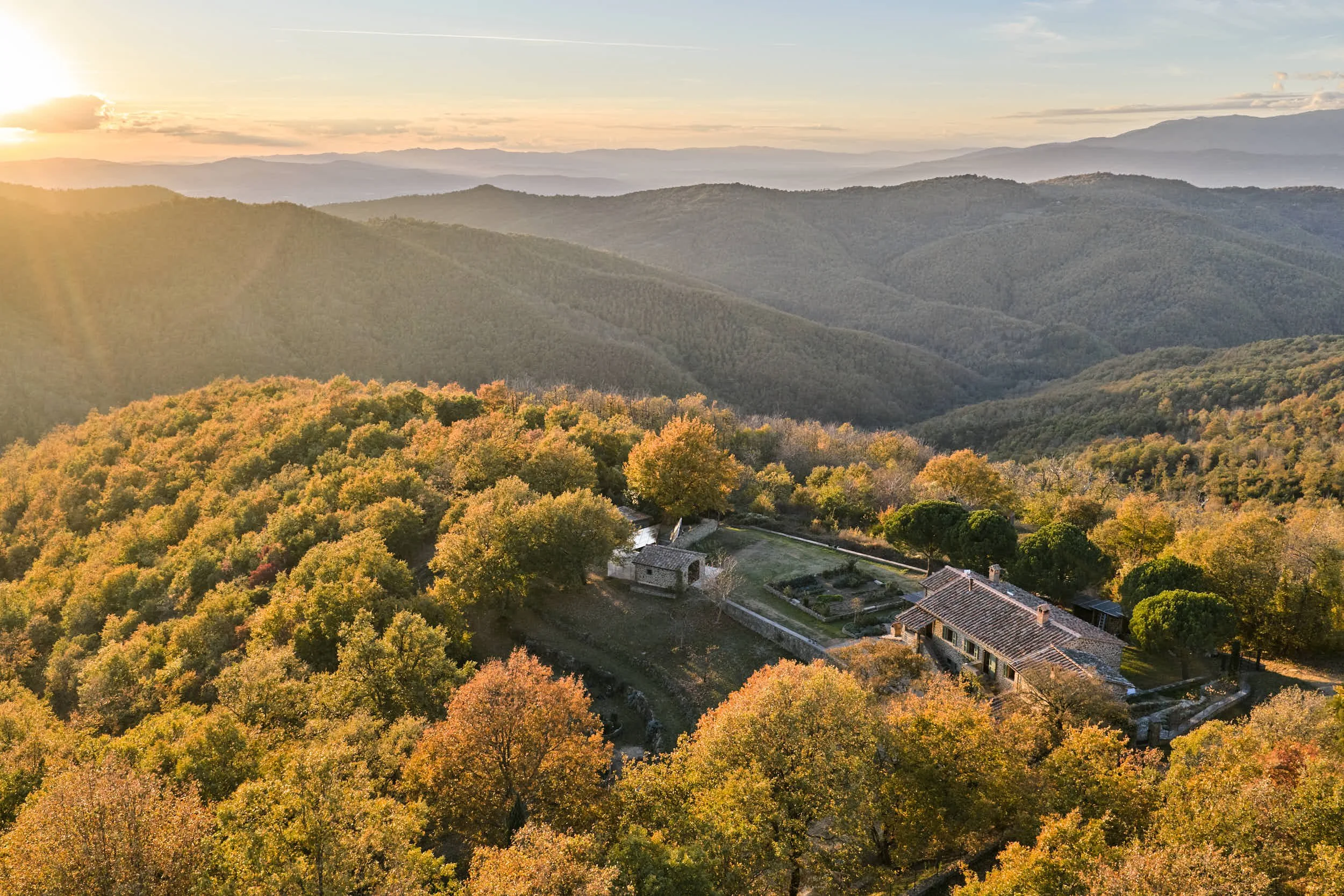 Landschaft mit Hügeln, Bäumen im Herbst, Sonnenuntergang und einem Haus in den Bäumen.