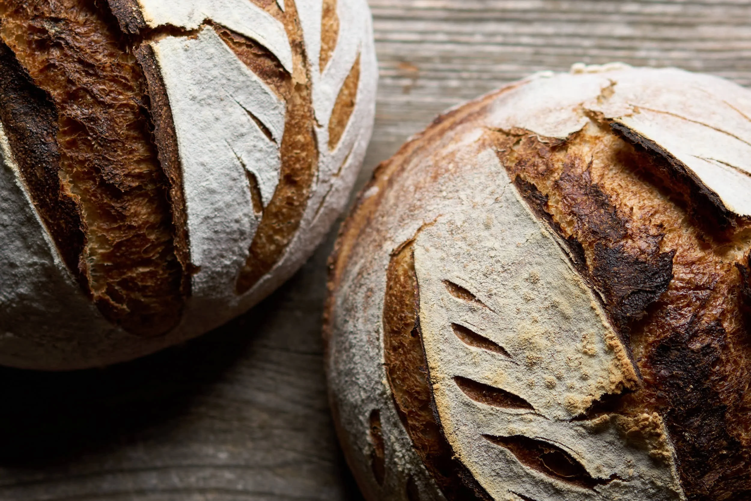 Zwei rustikale, gebäckartig gebackene Brote mit Mehl bestäubt, auf einem Holzbrett.