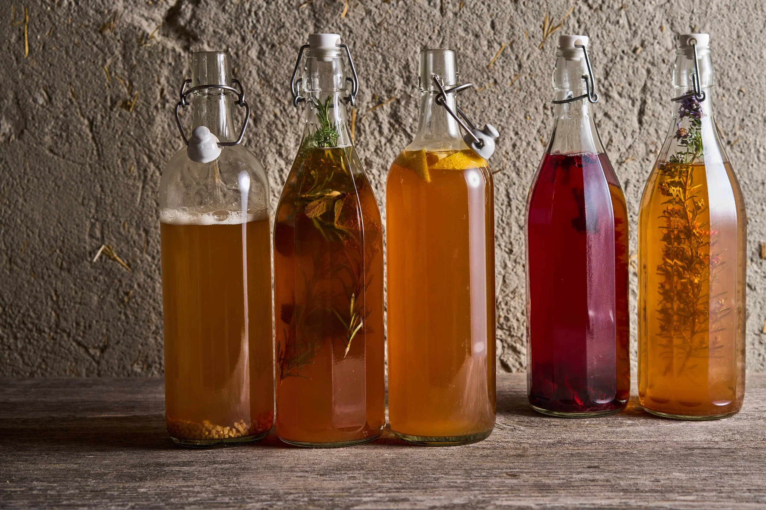 Fünf Glasflaschen mit verschiedenen gepressten Kräutern und Fruchtsäften auf einem Holztisch, vor einer steinernen Wand.