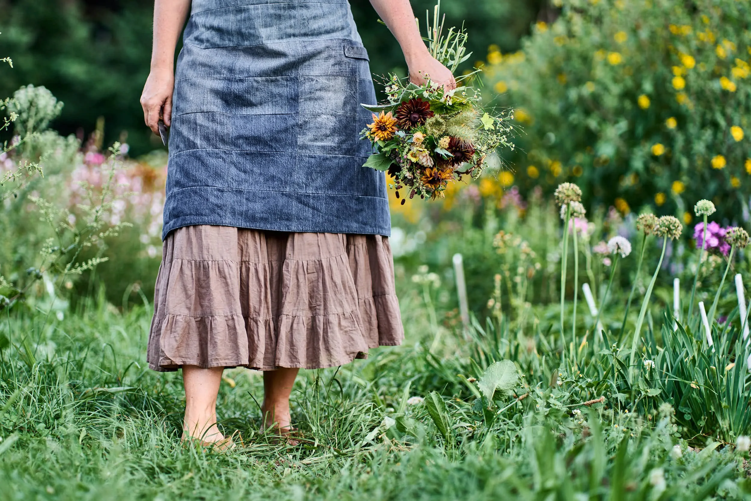 Frau in Rock und Schürze hält einen Blumenstrauß im Garten, voll mit bunten Blumen, in der Nähe von Gräsern und anderen Pflanzen.