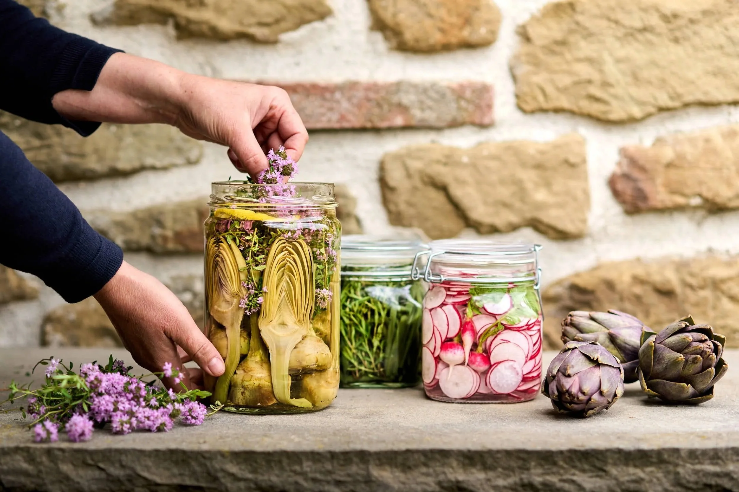 Hand, die eingelegte Ingwer, Radieschen und Kräuter in Gläser füllt, daneben frische Artischocken und ein Steinmauer im Hintergrund