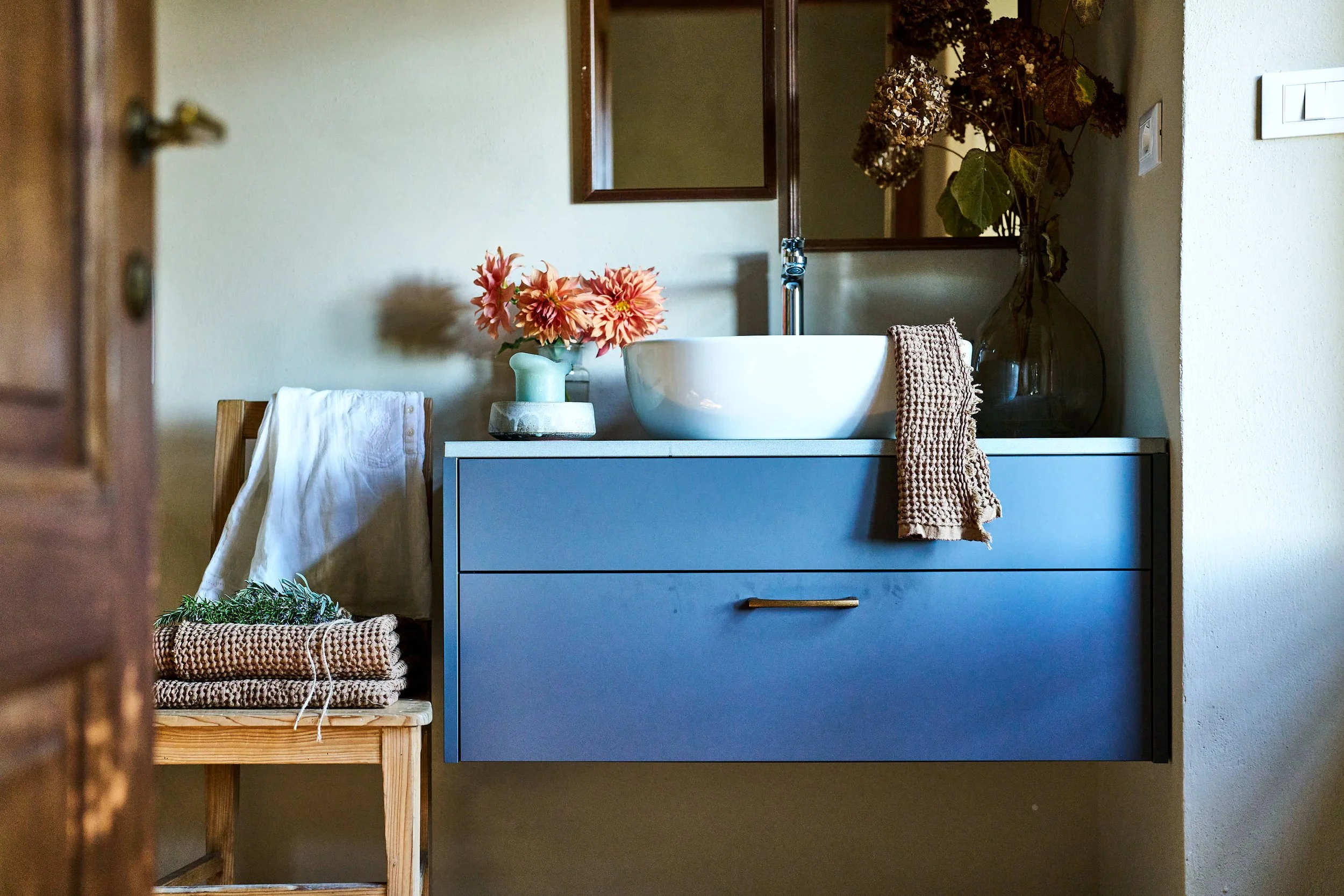Badezimmer mit blauer Waschtisch, einer weißen Waschschale, Lavendel und Blumen in Vasen, einem Handtuch und Spiegel.