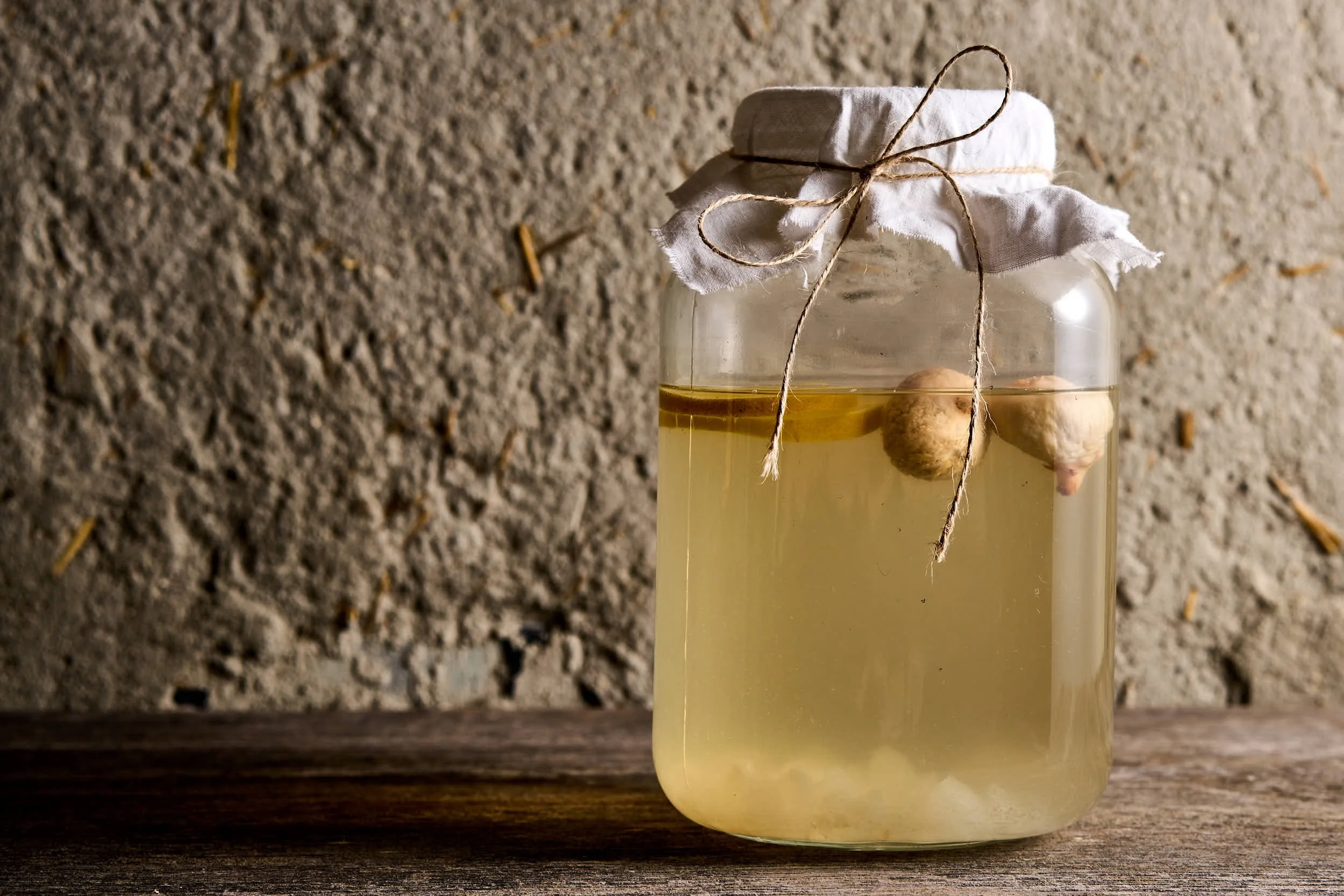 Ein Glas mit Zitronen-Ingwer-Hustensaft, abgedeckt mit einem Stofftuch, mit Ingwerstücken darin, auf einem Holzuntergrund vor einer Steinwand.