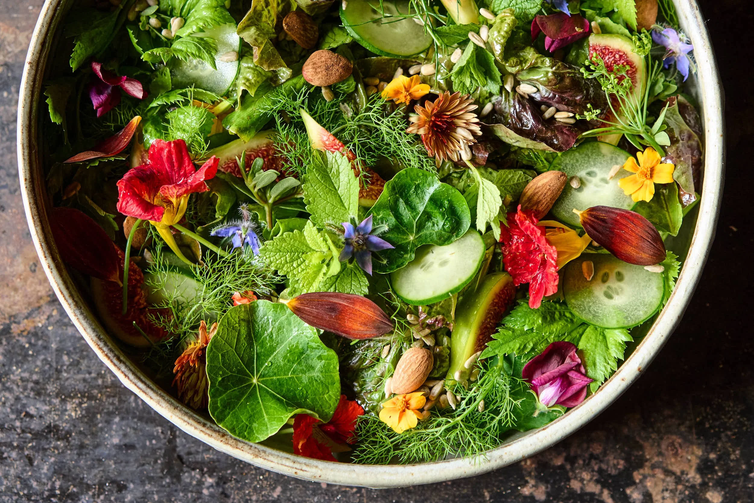 Bunter Salat mit grünen Blättern, Gurkenscheiben, essbaren Blumen, Samen und Nüssen in einer Schüssel, auf dunklem Untergrund